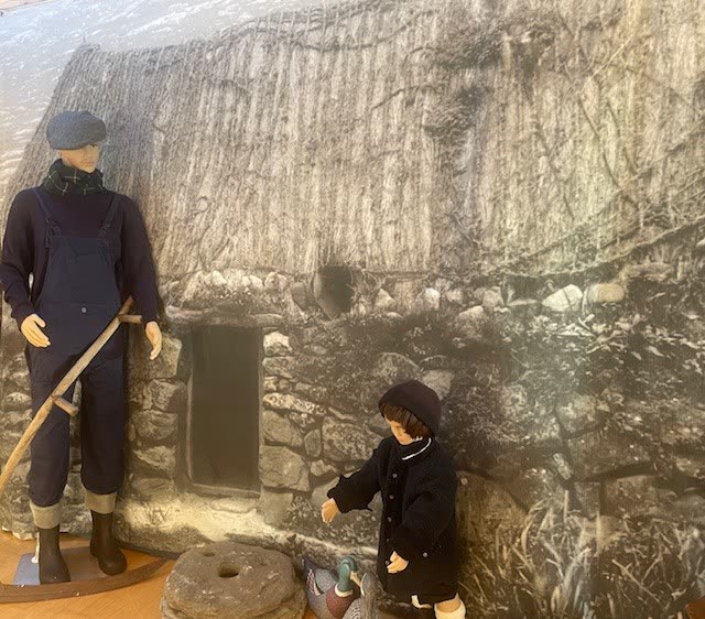 Figures of a man with a scythe and a child outside a thatched blackhouse